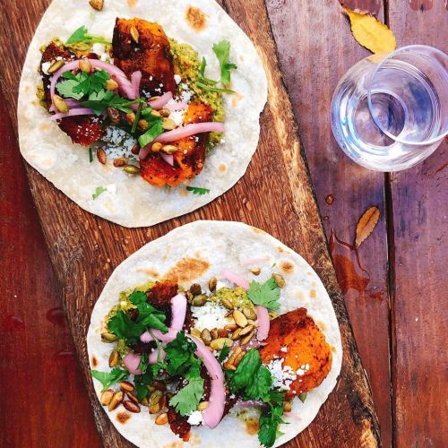 Two tacos on a wooden board, filled with roasted vegetables, guacamole, pickled onions, and garnished with cilantro and seeds.
