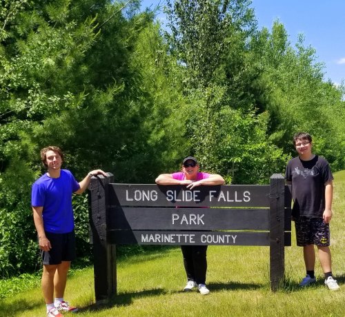 Three people stand by a sign for Long Slide Falls Park in Marinette County, surrounded by greenery on a sunny day.