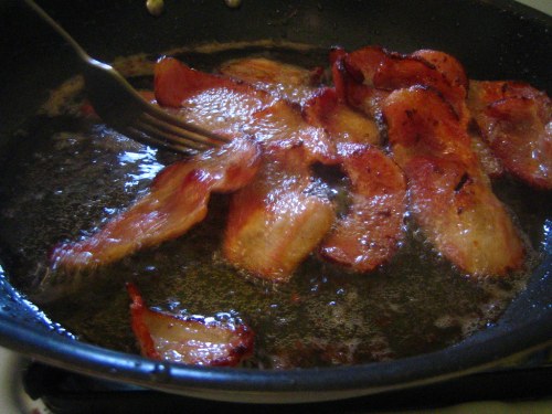 Sizzling bacon strips frying in a pan, with a fork lifting some pieces out of the hot oil.