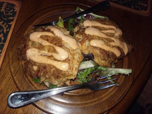 Two crab cakes on a bed of mixed greens, topped with a creamy sauce, served on a wooden plate with forks.
