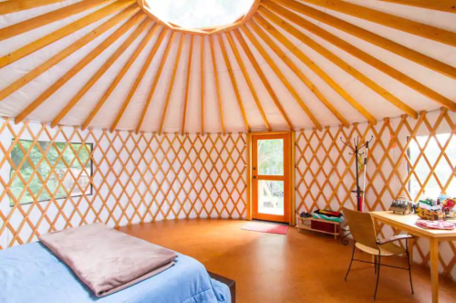 Interior of a yurt with wooden beams, a circular skylight, a bed, and a small table by the door.