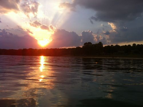 Sunset over a calm lake, with rays of light breaking through clouds and reflecting on the water's surface.