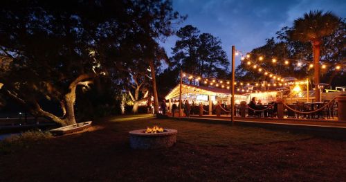 A cozy outdoor restaurant with string lights, surrounded by trees and a fire pit, set against a twilight sky.