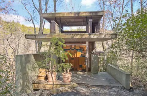 Modern two-story house nestled in a wooded area, featuring large windows and a wooden entrance surrounded by greenery.