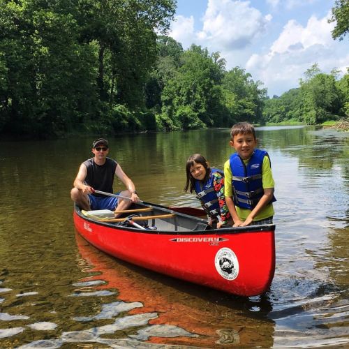 A man and two children paddle a red canoe on a calm river surrounded by lush greenery and blue skies.
