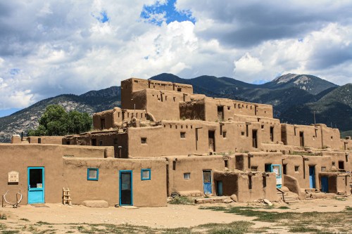 Adobe-style buildings with blue doors against a backdrop of mountains and cloudy skies.