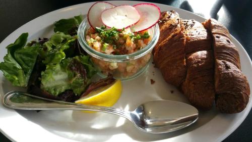 A plate with a croissant, salad, shrimp cocktail in a jar, radishes, and a lemon wedge, served with a spoon.