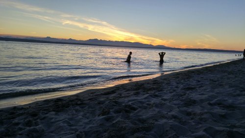 Silhouettes of two people wading in the water at sunset, with mountains in the background and a sandy beach in the foreground.