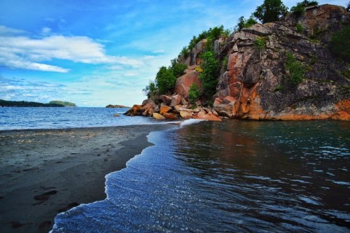 A serene shoreline with gentle waves lapping against a rocky cliff under a blue sky with scattered clouds.