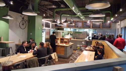 A bustling restaurant interior with diners at wooden tables, a visible kitchen, and industrial decor.