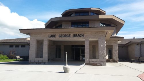 A modern building with a unique roof design, labeled "Lake George Beach," surrounded by a paved area and benches.