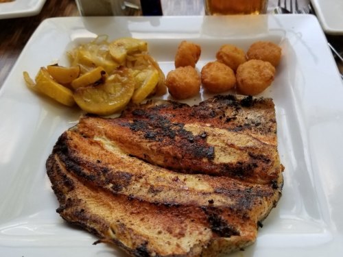 Grilled fish fillet served with sautéed yellow squash and golden hushpuppies on a white plate.