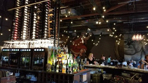 A stylish bar with a wine rack, colorful bottles, and warm lighting, featuring a mural and patrons enjoying the space.