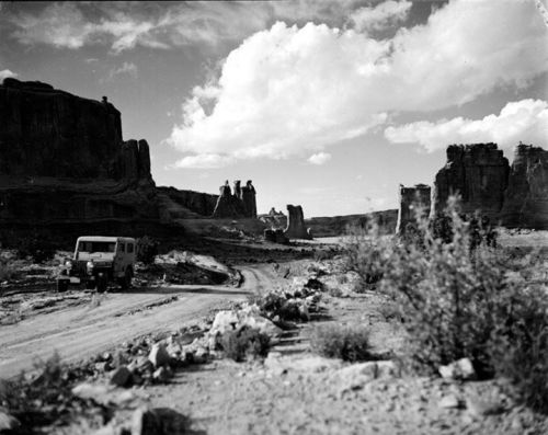 A vintage jeep on a dirt road surrounded by rocky formations and dramatic clouds in a desert landscape.