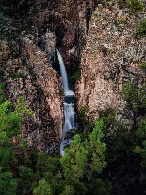 A waterfall cascades down rocky cliffs, surrounded by lush greenery and rugged terrain.