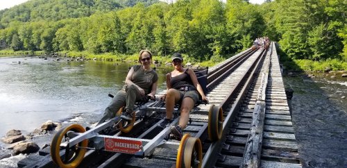 Two women sit on a rail bike on a wooden bridge over a river, surrounded by lush green trees and hills.