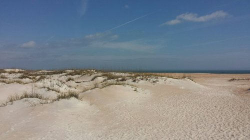 A serene beach scene with soft white sand, gentle dunes, and a clear blue sky above the calm ocean.