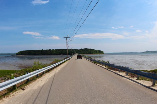 A narrow road stretches between water bodies under a clear blue sky, with power lines running alongside.