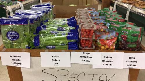 Display of fresh produce including romaine hearts, celery, grape tomatoes, Rainier cherries, and Bing cherries with prices.