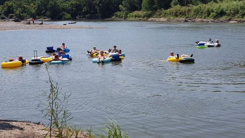 People relaxing on colorful inner tubes in a calm river, surrounded by greenery and a sandy bank.