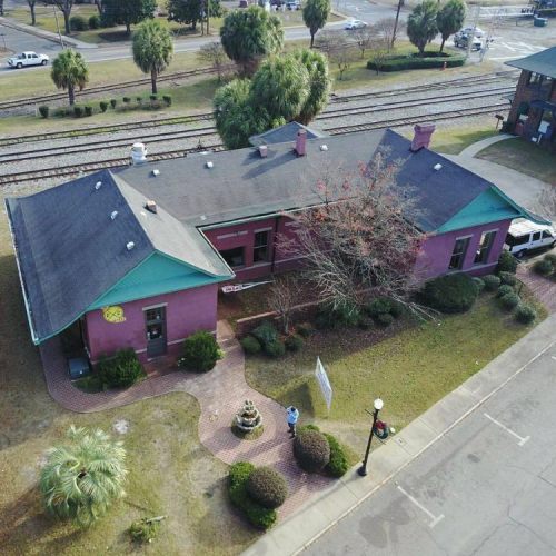 Aerial view of a purple building with a green roof, surrounded by trees and a lawn, near railway tracks.