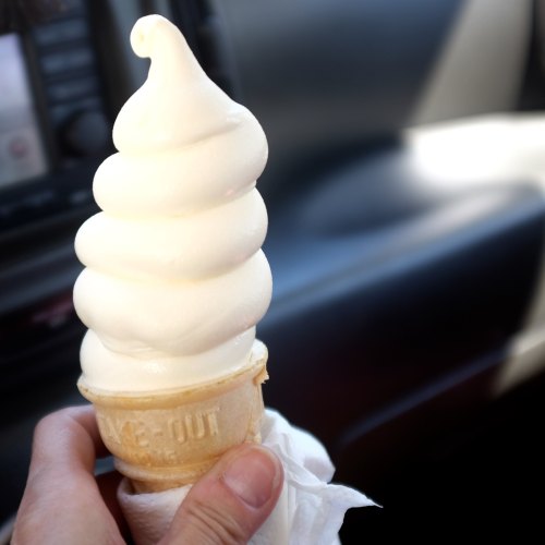 A hand holding a soft serve ice cream cone with a swirl of vanilla, set against a car interior background.