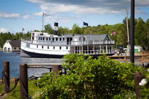A historic boat docked on a calm lake, surrounded by greenery and under a partly cloudy sky.