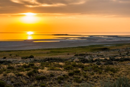 Sunset over a tranquil lake, with golden hues reflecting on the water and grassy shores in the foreground.