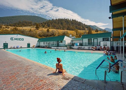 A woman sits by a large outdoor pool surrounded by mountains, with people enjoying the water and sun.