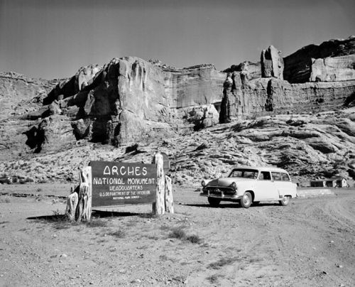 A vintage car parked near the Arches National Monument sign, surrounded by rocky terrain and cliffs.