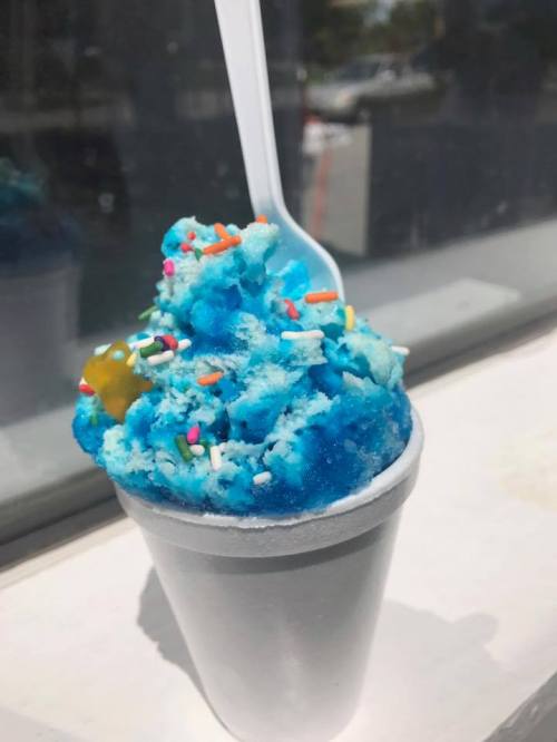 A cup of blue snow cone topped with colorful sprinkles and a gummy candy, sitting on a table.