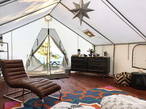 Cozy interior of a tent with a lounge chair, colorful rug, and wooden furniture, featuring a view of trees outside.
