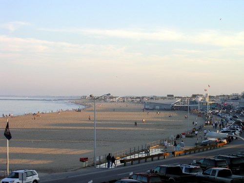A beach scene at sunset with people on the sand, cars parked nearby, and a calm ocean in the background.