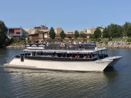 A large boat with passengers cruising on a calm river, surrounded by trees and buildings in the background.