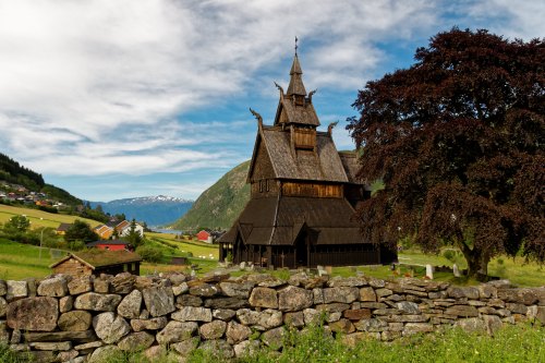 A wooden church with a tall spire stands beside a stone wall, surrounded by green hills and a blue sky.