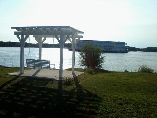 A white pergola with a swing overlooks a river, with a large boat in the background and greenery surrounding the area.