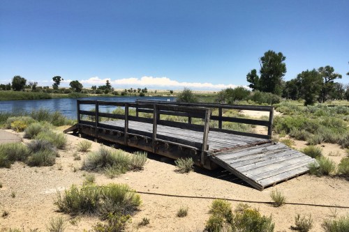 A weathered wooden dock on a sandy riverbank, surrounded by shrubs and trees under a clear blue sky.