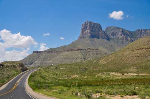 A winding road leads through green hills, with a prominent rocky mountain under a blue sky with scattered clouds.