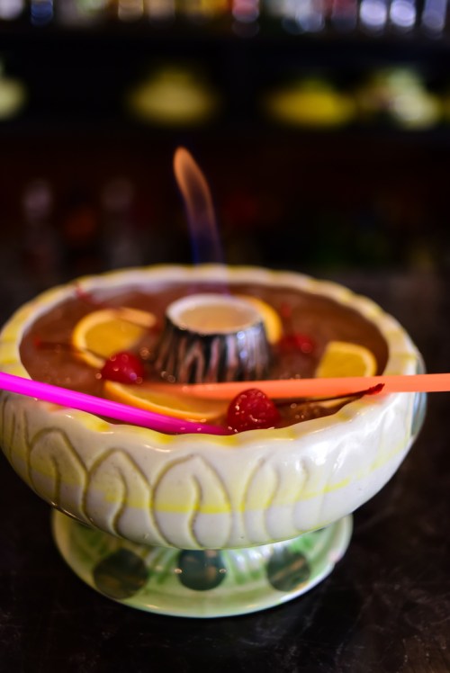 A colorful cocktail bowl with flaming liquid, garnished with lemon slices and raspberries, served with bright straws.