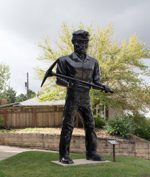 A large bronze statue of a miner holding a pickaxe, set against a backdrop of trees and a cloudy sky.