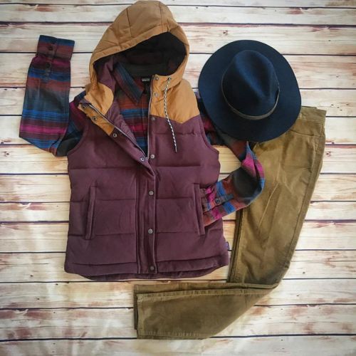A stylish outfit featuring a maroon puffer vest, plaid shirt, brown pants, and a black hat on a wooden background.