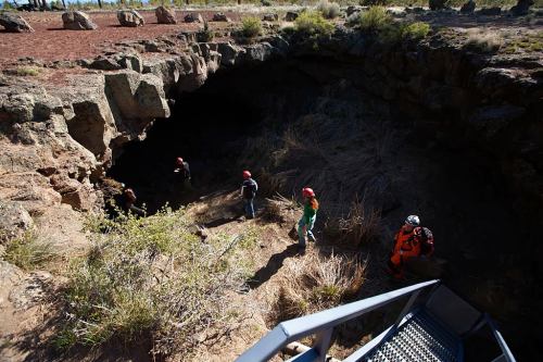 A group of people in helmets exploring a cave entrance surrounded by rocky terrain and sparse vegetation.
