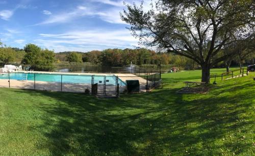 A serene outdoor scene featuring a fenced swimming pool beside a lake, surrounded by trees and green grass.