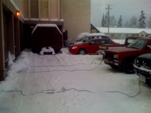 Snow-covered parking area with two vehicles and power cords running to them, indicating cold weather preparations.