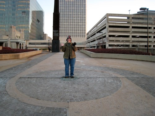 A person stands on a circular stone pattern in an urban area, surrounded by buildings and a parking garage.