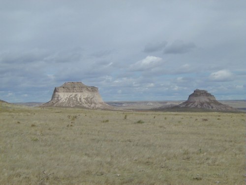 Two prominent buttes rise from a grassy plain under a cloudy sky, showcasing a rugged, natural landscape.