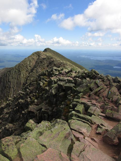 A rocky mountain peak under a blue sky with clouds, overlooking a vast landscape and distant water bodies.