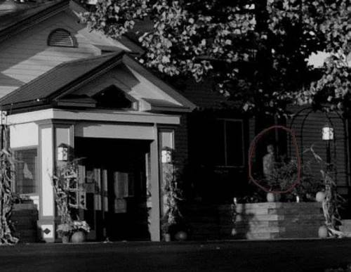 A dimly lit house exterior with a figure partially visible near the entrance, surrounded by autumn decorations.