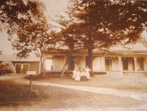 A sepia-toned photo of a large house with four children sitting on the lawn under a tree.
