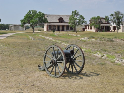 A historic cannon on grassy land with two buildings and trees in the background under a clear sky.